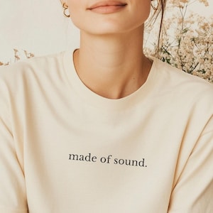 Könnte beinhalten: Cremefarbenes T-Shirt mit dem Text "made of sound" in schwarzer, minimalistischer Schrift. Das Model trägt kleine goldene Creolen und hat die Haare zu einem Dutt hochgesteckt. Der Hintergrund umfasst Trockenblumen und einen Stoff in neutralen Tönen.