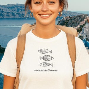 May include: A white short-sleeved t-shirt featuring a graphic of three stacked fish illustrations in black ink. Below the fish, the text reads Modulate to Summer in a simple black font. The shirt has a crew neckline and a standard fit.