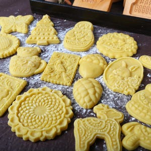 Puede incluir: Una colecci&oacute;n de galletas sin hornear, de color amarillo, en varias formas, incluyendo una mariposa, un &aacute;rbol de Navidad y un girasol. Las galletas est&aacute;n dispuestas sobre una superficie oscura espolvoreada con polvo blanco, sugiriendo una preparaci&oacute;n para hornear.