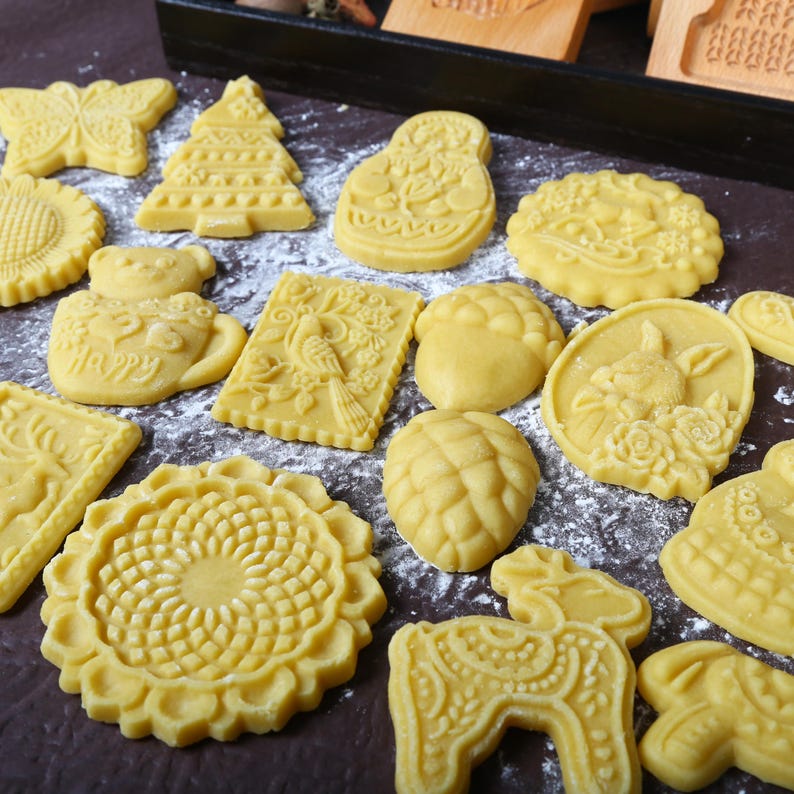 Puede incluir: Una colecci&oacute;n de galletas sin hornear, de color amarillo, en varias formas, incluyendo una mariposa, un &aacute;rbol de Navidad y un girasol. Las galletas est&aacute;n dispuestas sobre una superficie oscura espolvoreada con polvo blanco, sugiriendo una preparaci&oacute;n para hornear.