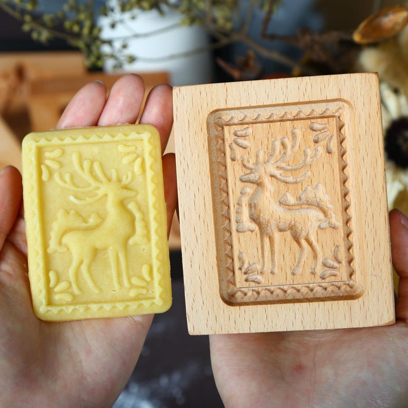 Puede incluir: Un molde de galletas de madera y una galleta prensada. El molde es rectangular con un dise&ntilde;o de ciervo tallado. La galleta es amarilla y tiene el mismo dise&ntilde;o de ciervo. El molde es de madera clara.