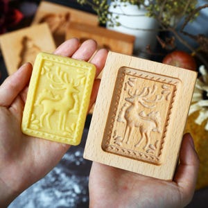 Puede incluir: Un molde de galletas de madera y una galleta prensada, ambos con un dise&ntilde;o detallado de ciervo. La galleta es rectangular con un tono amarillo, mientras que el molde es de madera clara. El dise&ntilde;o del ciervo est&aacute; enmarcado por un borde decorativo.