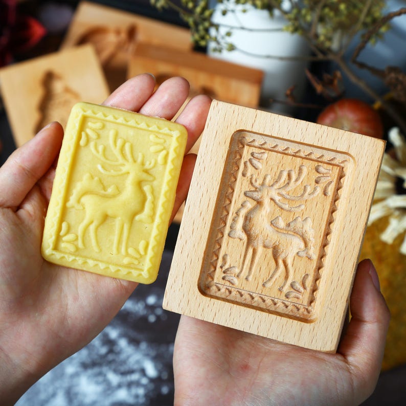 Puede incluir: Un molde de galletas de madera y una galleta prensada, ambos con un dise&ntilde;o detallado de ciervo. La galleta es rectangular con un tono amarillo, mientras que el molde es de madera clara. El dise&ntilde;o del ciervo est&aacute; enmarcado por un borde decorativo.