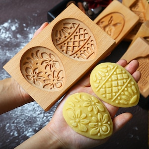 May include: Wooden egg-shaped cookie molds with floral and geometric designs. Two molded cookies are held in a hand, showcasing the detailed patterns. The molds are made of light-colored wood, perfect for Easter baking.
