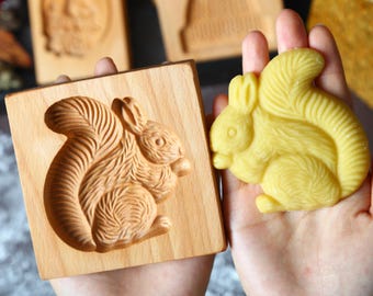 May include: A wooden squirrel-shaped cookie mold next to a yellow squirrel-shaped treat. The mold is square and features a detailed carving of a squirrel. Other wooden molds with animal designs are in the background.