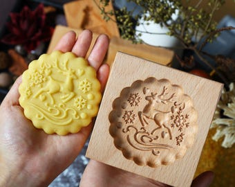 May include: A round, yellow cookie with a deer and snowflake design is held next to a wooden cookie mold with the same design. The mold is square with a scalloped edge. The cookie has a scalloped edge as well.