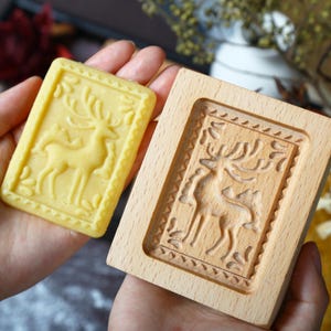 Puede incluir: Una galleta rectangular de color amarillo claro con un dise&ntilde;o detallado de ciervo se sostiene junto a un molde de galletas de madera con el mismo dise&ntilde;o de ciervo. La galleta tiene un borde decorativo. El molde de madera es de madera clara.