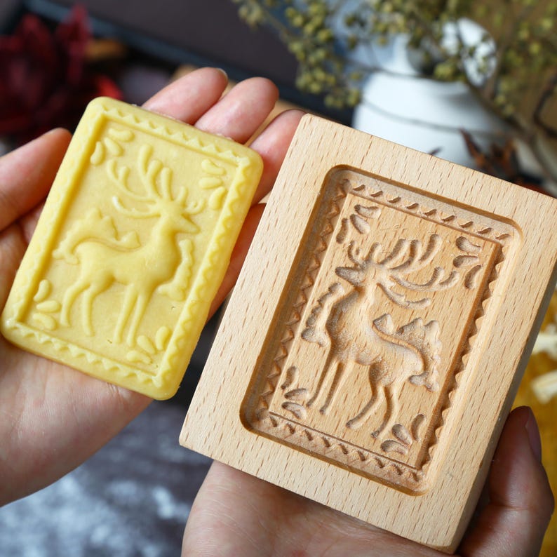 Puede incluir: Una galleta rectangular de color amarillo claro con un dise&ntilde;o detallado de ciervo se sostiene junto a un molde de galletas de madera con el mismo dise&ntilde;o de ciervo. La galleta tiene un borde decorativo. El molde de madera es de madera clara.
