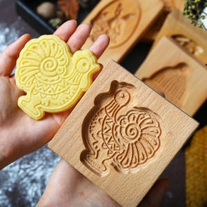 May include: A yellow, rooster-shaped cookie next to a wooden cookie mold with a rooster design. The cookie and mold are held in a person's hands. Other wooden molds with animal designs are in the background.