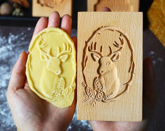 May include: A wooden cookie stamp with a carved deer design, held next to a cookie with the same embossed image. The stamp features a deer within a circular frame with pinecones and foliage. The cookie is a light yellow color.