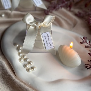 Może przedstawiać: Biała świeca w kształcie muszli z płonącym płomieniem spoczywa na białej, organicznie ukształtowanej tacy. Obecne jest również małe pudełko upominkowe z kremową wstążką i etykietą z napisem "Muhanad & Nira, June 28th 2016", wraz z rozrzuconymi perłami.