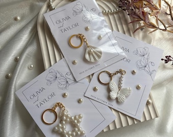 May include: Keychains with gold rings and pearl embellishments are displayed on cards that read "OLIVIA and TAYLOR" and "30TH OCTOBER 2026". The keychains feature a star, a butterfly, and a shell design. Scattered pearls and floral accents complete the presentation.