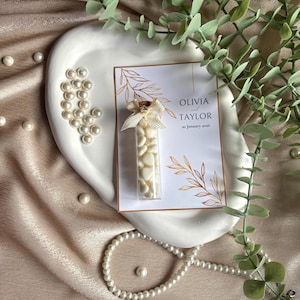 Puede incluir: Un plato de cerámica blanca contiene un pequeño tubo de vidrio lleno de caramelos blancos, una tarjeta con los nombres "Olivia Taylor" y la fecha, y un lazo decorativo. Perlas dispersas y un collar de perlas rodean el plato, con follaje verde en el fondo.