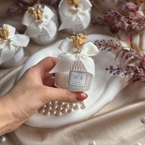 Op de afbeelding: Transparante, cilindrische kaarsbedankjes met witte strikken en gedroogde bloemaccenten. Elk bedankje heeft een klein label met de initialen "D B" en de woorden "Wedding Journey". Verschillende bedankjes zijn gerangschikt op een wit bord.