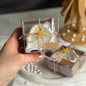May include: A square clear box containing a white shell-shaped candle, wrapped in brown twine with a small yellow starfish accent. A rectangular kraft paper tag is attached, featuring the printed text Hailey & James 20.06.2024.