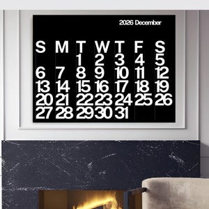 May include: A framed black and white calendar for December 2026. The calendar displays the days of the week across the top and numbered dates below. The calendar is mounted above a fireplace with a lit fire and a beige armchair.