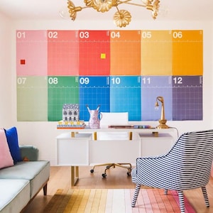 Puede incluir: Un espacio de oficina colorido con un gran calendario de colores del arcoíris en la pared. La habitación cuenta con un escritorio blanco, una silla a rayas y una alfombra degradada. Una lámpara de araña dorada cuelga del techo y se ve un sofá con cojines.