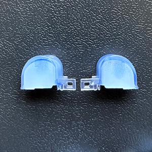 May include: Two translucent blue plastic components, each featuring a semi-circular dome shape and a small rectangular tab with a circular hole on the side. These identical parts are positioned side-by-side, showcasing a smooth, curved surface.