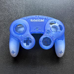 Puede incluir: Una carcasa de mando Nintendo GameCube en azul translúcido con un efecto degradado que se aclara hacia la parte inferior de las empuñaduras. La parte frontal muestra el logotipo y texto Nintendo GameCube en blanco, con huecos para botones.