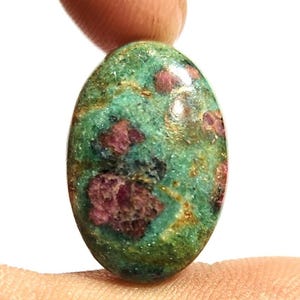 Pode incluir: Gema ovalada com base verde e manchas vermelhas. A pedra polida é segurada entre os dedos. Provavelmente é um rubi em zoisite. A pedra tem aproximadamente 2,5 cm de comprimento.