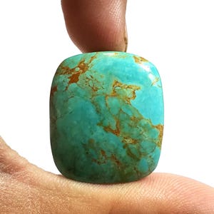 Könnte beinhalten: Ein polierter, rechteckiger Türkis-Edelstein mit braunen Adern. Der Stein wird zwischen zwei Fingern vor einem weißen Hintergrund gehalten. Die Türkisfarbe ist ein leuchtendes Blaugrün.