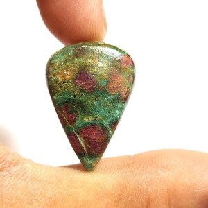 Pode incluir: Uma pedra preciosa em forma de gota com uma mistura de cores. A pedra apresenta uma base verde e dourada com manchas vermelhas e castanhas. A pedra preciosa é segurada entre dois dedos contra um fundo branco.