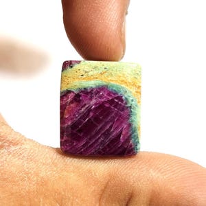 Puede incluir: Una gema rectangular con un centro magenta intenso, bordeada de verde claro y beige. La piedra se sostiene entre los dedos, resaltando su textura natural. Esta gema probablemente se utiliza en joyería.