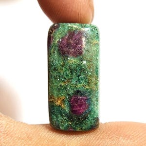 Puede incluir: Una gema rectangular con una base verde e inclusiones rojas. La piedra se sostiene entre dos dedos sobre un fondo blanco. La gema tiene una superficie pulida y una apariencia natural y orgánica.