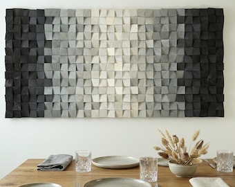 Op de afbeelding: Een rechthoekig wandkunstwerk bestaande uit talloze kleine, vierkante houten blokjes gerangschikt in een raster. De blokjes hebben een kleurverloop, variërend van diepzwart aan de buitenranden tot lichtgrijs en wit in het midden.