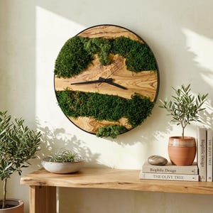 May include: A round wall clock featuring a natural wood grain face with dark green moss accents and thin black hands. The clock is encased in a slim black circular frame, showcasing a blend of organic wood textures and vibrant green preserved moss.