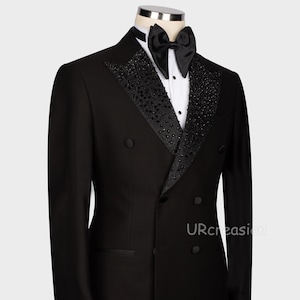 May include: A formal black double-breasted tuxedo jacket with a satin lapel and a black bow tie. The lapel is embellished with black beads. The jacket has a white dress shirt underneath. The text "URcreasion" is visible on the right side of the jacket.