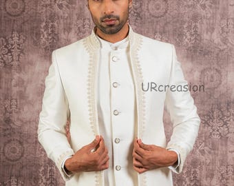 May include: Ivory-colored formal suit with intricate embroidery along the jacket's edges and collar. The suit includes a matching vest and trousers. The jacket is open, revealing the vest and button details. The background is a patterned, muted brown.