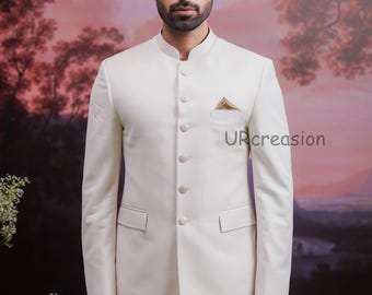 May include: A cream-colored suit with a mandarin collar, featuring a pocket square and button-up closure. The suit is tailored and worn by a person. The background is a painted landscape with trees and a sunset.