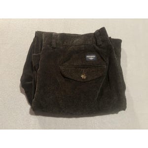 Puede incluir: Pantalones de pana de color marrón oscuro con tejido de canalé texturizado, un bolsillo trasero con botón y una etiqueta rectangular azul marino con el texto blanco DOCKERS cosido sobre el bolsillo. Los pantalones incluyen trabillas.