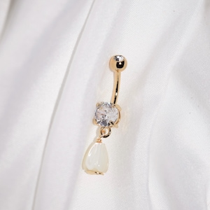 May include: A gold-colored belly button ring with a clear, round gemstone at the top and a larger, clear, round gemstone in the center. A pear-shaped, white pendant hangs from the center gemstone. The ring is set against a white background.