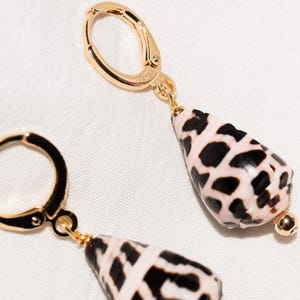 May include: Gold-tone hoop earrings with a dangling, teardrop-shaped shell pendant. The shell is white with black spots, and the earrings have a small gold bead at the bottom of the pendant. The earrings are displayed on a white surface.