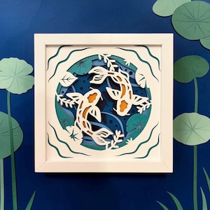 May include: A square white frame containing a multi-layered paper art piece featuring two koi fish in shades of white and orange. The fish are surrounded by teal and blue water layers with cut-out lily pads and wavy patterns in a circular design.