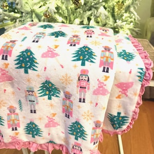 May include: A white blanket with a pink ruffle trim, featuring a festive pattern of Christmas trees, nutcrackers, and ballerinas in pink, teal, and gold. The blanket is folded over a white metal stand.