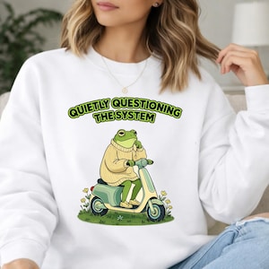 May include: White sweatshirt with a cartoon frog on a scooter. The frog is in a beige sweater, riding a mint green scooter. The text "QUIETLY QUESTIONING THE SYSTEM" is above the frog in a green font.