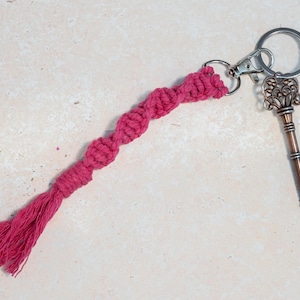 May include: A hot pink macramé keyring featuring a spiral knot pattern that ends in a short tassel. The cord is attached to a silver-toned metal lobster clasp and a split ring, which also holds a vintage-style copper-coloured skeleton key.