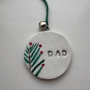 Könnte beinhalten: Weißer Keramik-Weihnachtsschmuck mit dem geprägten Wort "DAD". Er hat ein bemaltes grünes und rotes Design, das an einer grünen Schnur und einer silbernen Glocke befestigt ist.