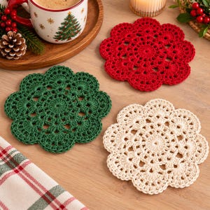 May include: Three round crochet coasters in red, dark green, and cream colors. Each coaster features a scalloped edge and an intricate, openwork floral pattern. The coasters are arranged on a wooden surface alongside a mug and a plaid cloth.