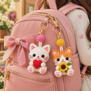 May include: Two crocheted cat keychains attached to a pink backpack. One white cat holds a red heart, and one orange tabby cat holds a yellow sunflower. Both cats have pink cheeks, black eyes, and pink paw pads, suspended by gold-toned metal clasps.