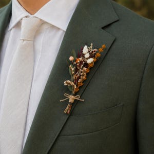 Könnte beinhalten: Dunkelgrüner Blazer mit weißem Hemd und hellbeiger Krawatte. Ein kleiner Strauß Trockenblumen in Orange, Weiß und Burgunderrot ist an der Revers befestigt und mit Bindfaden gebunden.