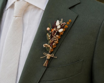 Può includere: Una giacca verde scuro con una camicia bianca e una cravatta beige chiaro. Un piccolo bouquet di fiori secchi arancioni, bianchi e bordeaux è appuntato al bavero, legato con spago.