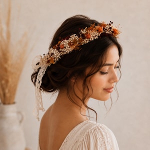 Op de afbeelding: Een bloemenkrans met gedroogde bloemen in oranje, bordeauxrode en crèmetinten. De krans wordt gedragen op donker haar en heeft kanten banden. De stijl is bohemien en natuurlijk, geschikt voor bruiloften of speciale gelegenheden. Een decoratief haaraccessoire.