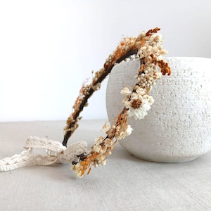 Op de afbeelding: Een delicate bloemenkrans met gedroogde bloemen in crème, bruin en oranje tinten. De krans is bevestigd aan een donkerbruine band en heeft een crèmekleurige kanten strik. De krans wordt naast een witte, getextureerde vaas geplaatst.