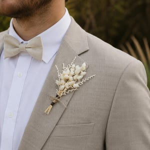 Könnte beinhalten: Ein beigefarbener Leinenanzug mit passender Fliege und weißem Hemd. Ein kleiner Strauß getrockneter weißer Blumen ist an der Anzugjacke befestigt. Der Anzug hat eine hellbraune Farbe.