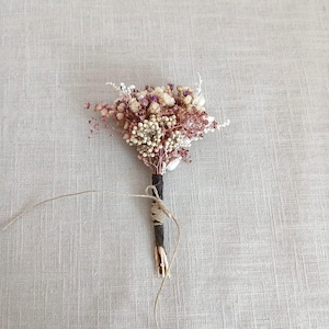 Può includere: Un piccolo bouquet di fiori secchi con uno stelo marrone scuro avvolto nello spago. Il bouquet presenta un mix di fiori bianchi, rosa e viola. Un pezzo di spago si estende dallo stelo. Lo sfondo è un tessuto beige chiaro.