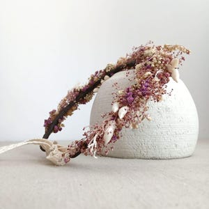 Può includere: Una corona floreale con fiori secchi nei toni del viola, bianco e beige. La corona è posta su una base marrone scuro e include un nastro color crema. La composizione floreale è delicata e naturale, perfetta per un'occasione speciale.